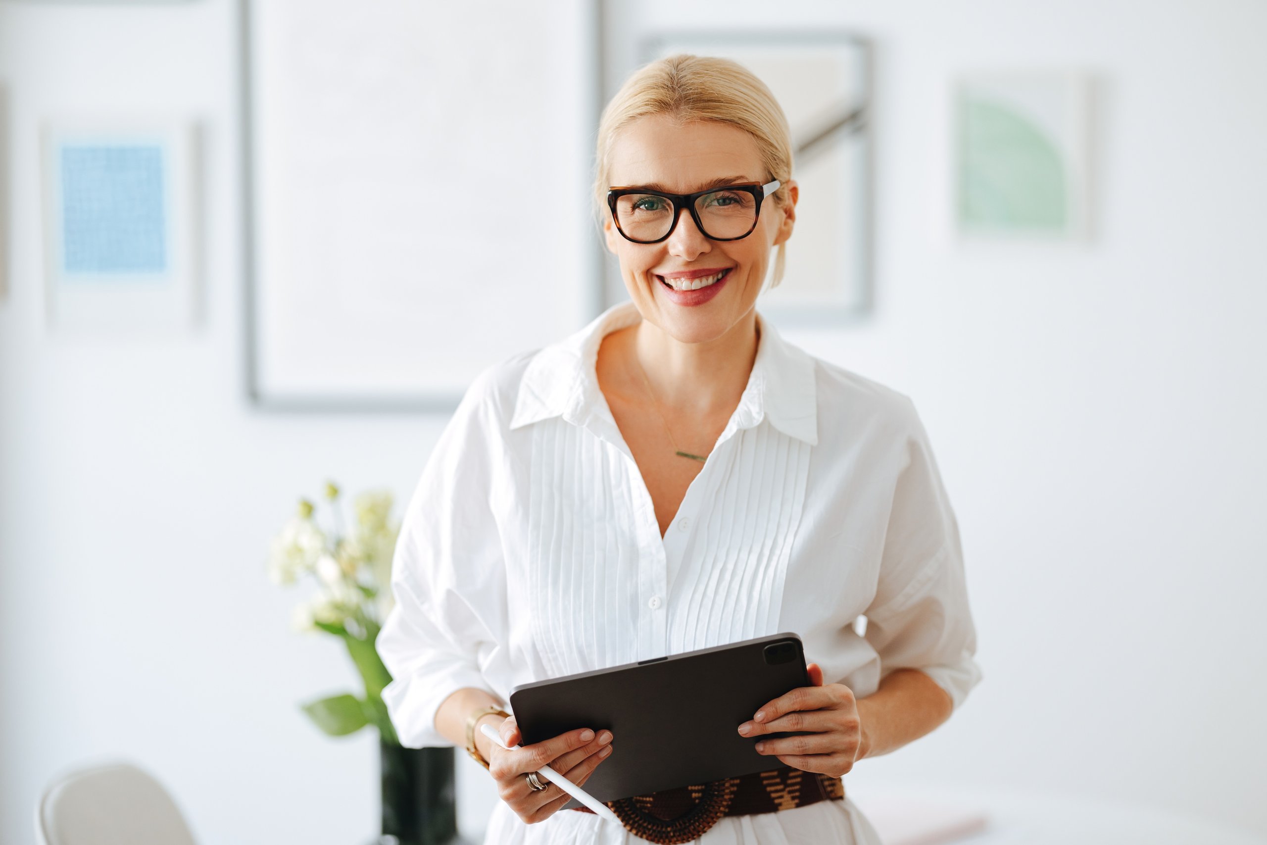 Blonde woman in glasses holds a tablet, smiling.