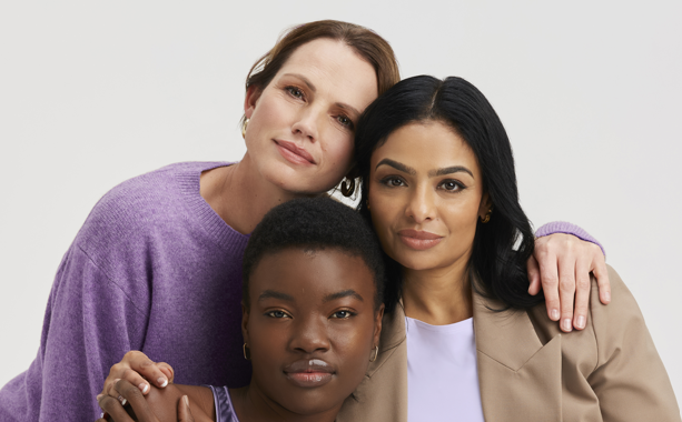 Three women in purple and beige attire, embracing, smiling.