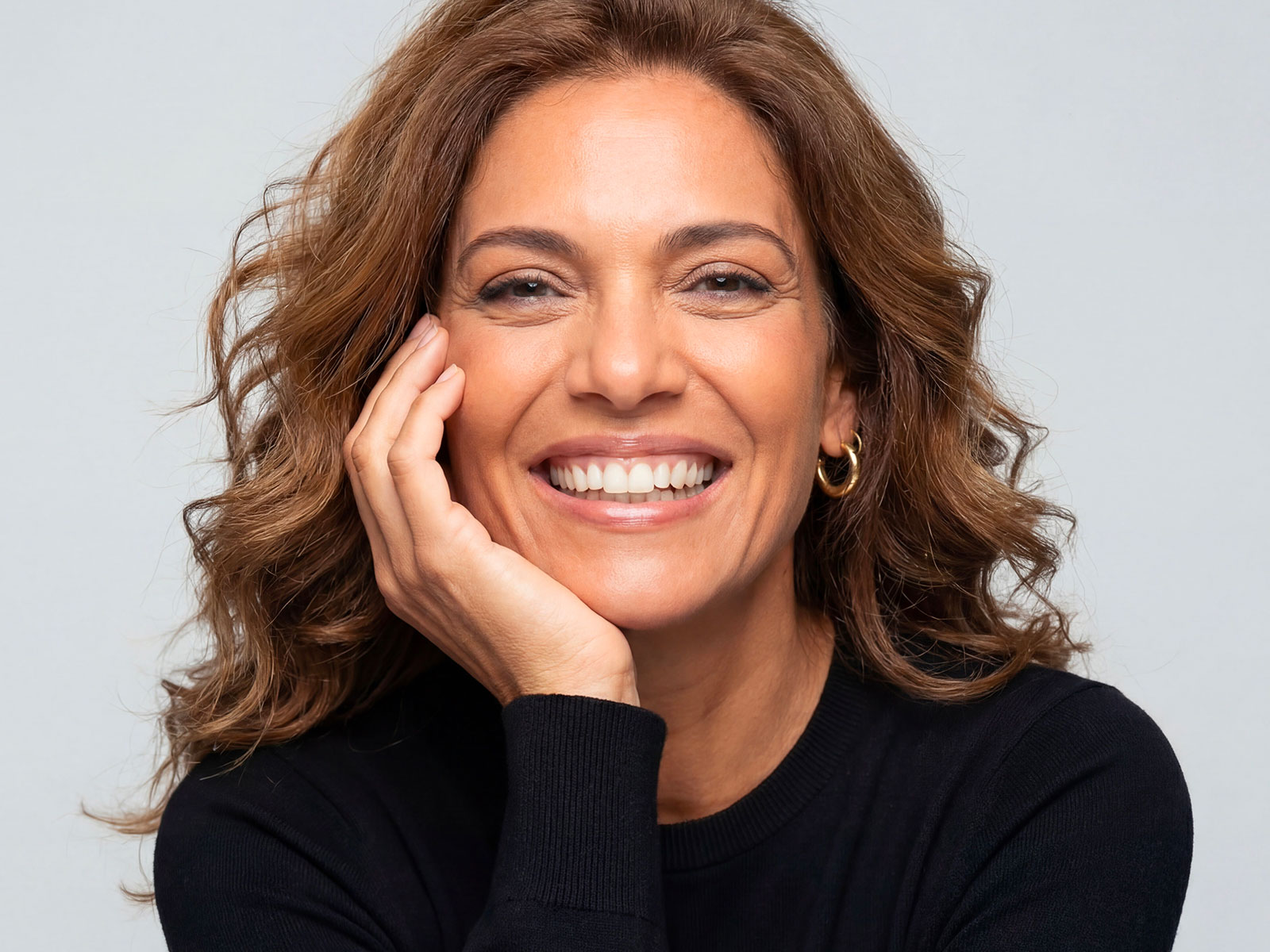 A woman with brown wavy hair, gold hoop earrings, and a black sweater smiles with her hand on her chin.