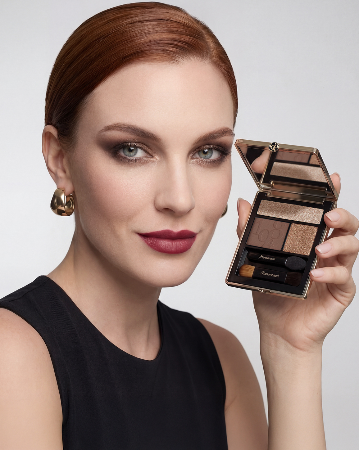 Woman with red hair and bold makeup holds an open Marionnaud eyeshadow palette with four shades and two applicators against a white background.