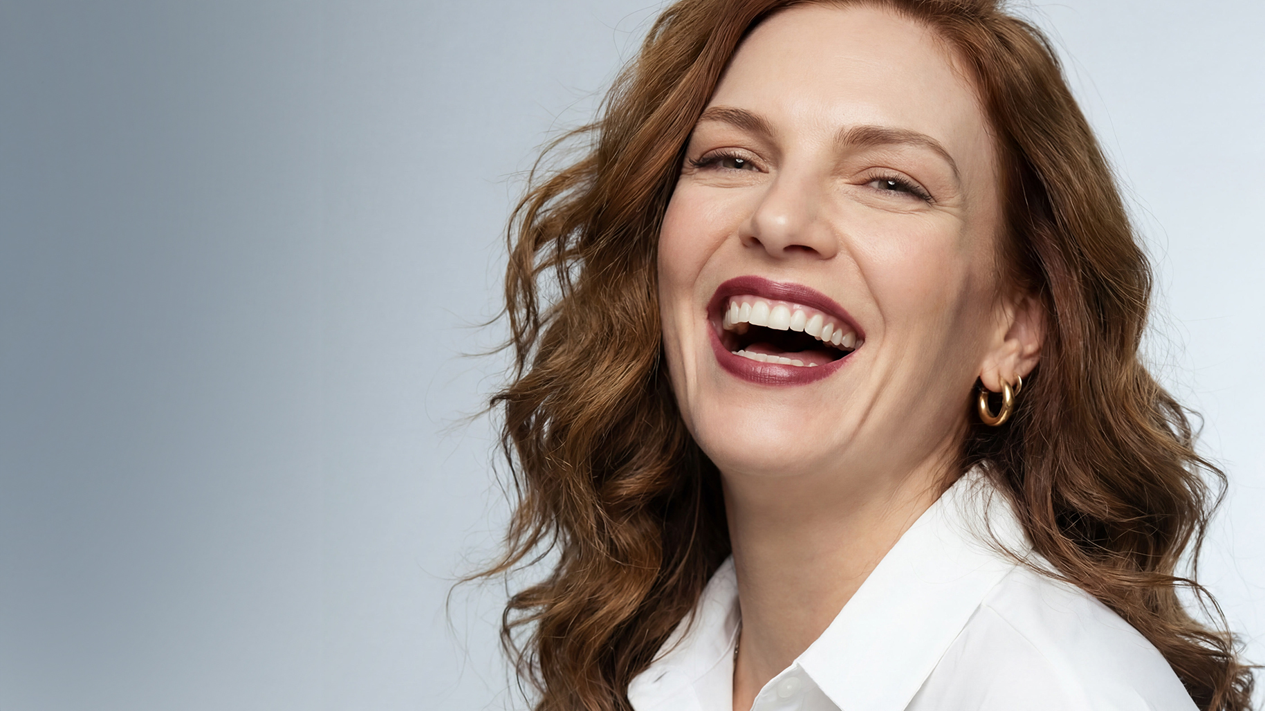 A woman with auburn hair laughs wholeheartedly, her mouth open and eyes crinkled. She wears a white collared shirt and gold hoop earrings against a gray background.