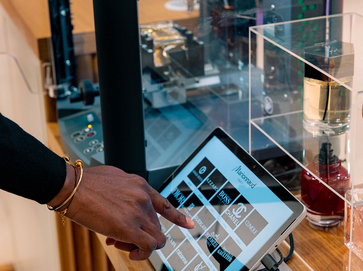 A person's hand, adorned with a gold bracelet, touches a tablet displaying brand logos for perfumes like Dior, Chanel, and Hermès.